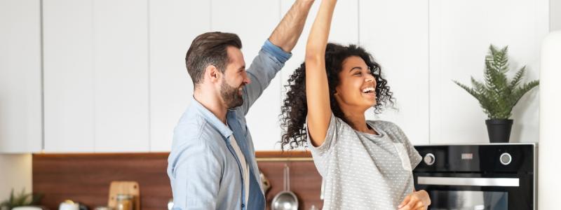 Een lachend stel danst samen in de keuken, terwijl ze genieten van muziek en een vrolijk moment.